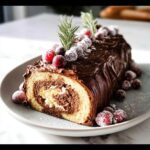 A slice of Classic Buche de Noël (Yule Log) cake with rich chocolate frosting, rolled sponge, and sugared cranberries.
