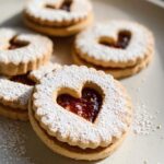 Close-up of Gluten-Free Linzer Cookies dusted with powdered sugar, filled with red jam and a heart-shaped cutout.
