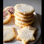 A stack of freshly baked gluten-free sugar cookie cutouts dusted with powdered sugar, with star-shaped and sprinkle-covered cookies nearby.