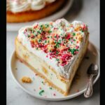 A generous slice of Sugar Cookie Cheesecake topped with whipped cream, colorful sprinkles, and cookie crumbles.
